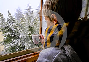 Blonde boy with scarf looking out window and holding in hands mug with hot chocolate across winter view