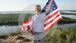 Blonde boy waving national USA flag outdoors over blue sky at the river bank