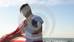 Blonde boy waving national USA flag outdoors over blue sky at the river bank