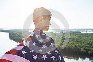 Blonde boy waving national USA flag outdoors over blue sky at the river bank