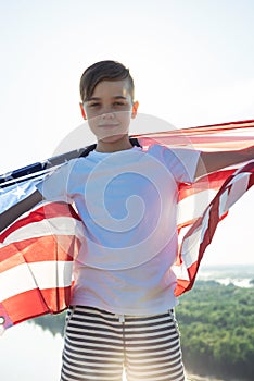 Blonde boy waving national USA flag outdoors over blue sky at the river bank