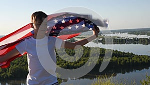 Blonde boy waving national USA flag outdoors over blue sky at the river bank