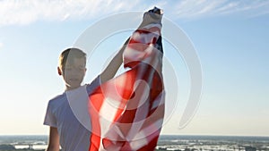 Blonde boy waving national USA flag outdoors over blue sky at the river bank