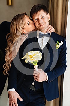 the blonde bride gently hugs the groom's shoulders by the window.