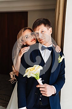 the blonde bride gently hugs the groom's shoulders by the window.