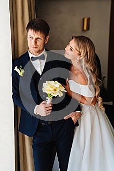 the blonde bride gently hugs the groom's shoulders by the window.