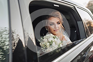 A blonde bride, smiling sincerely, sits in a black car on her wedding day