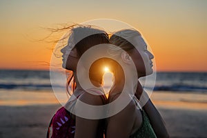 Blonde and brunette girlfriends are clinging back to each other against sunset over the sea. Happy lesbian european couple is