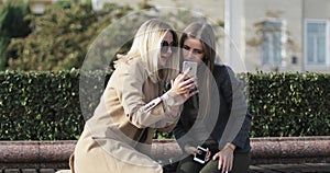 Blonde and brunette looking at mobile phone screen smiling and laughing sitting on bench in park.
