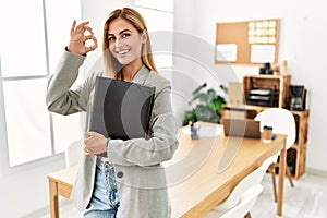 Blonde business woman at the office smiling positive doing ok sign with hand and fingers