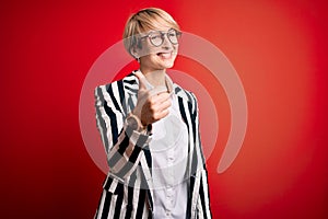 Blonde business woman with short hair wearing glasses and striped jacket over red background doing happy thumbs up gesture with