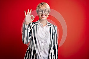 Blonde business woman with short hair wearing glasses and striped jacket over red background smiling positive doing ok sign with