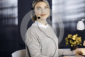 Blonde business woman talking by headset while sitting in modern call center office. Telemarketing and customer service