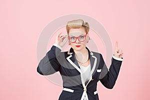 Blonde businesswoman with red glasses pointing up by finger on pink background