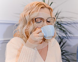 Blonde casual closed woman drink tea. Female with glasses hold a small light blue mug with hot coffee.