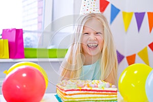 Blonde caucasian girl laughing at camera near birthday rainbow cake. Festive colorful background with balloons