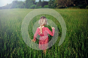 Blonde Caucasian girl in pink rustic dress walking in tall grass on meadow at sunset