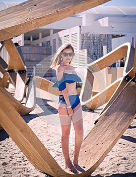 Blonde Caucasian Woman on the Beach With Blue Flounce Top  Swimsuit, Sitting on a Wood Structure and Looking Away photo