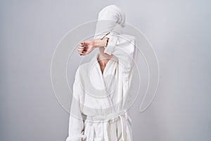 Blonde caucasian woman wearing bathrobe covering eyes with arm, looking serious and sad