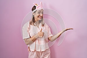 Blonde caucasian woman wearing sleep mask and pajama showing palm hand and doing ok gesture with thumbs up, smiling happy and