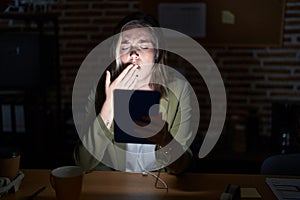 Blonde caucasian woman working at the office at night bored yawning tired covering mouth with hand