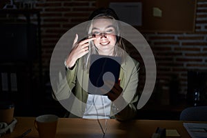 Blonde caucasian woman working at the office at night pointing with hand finger to face and nose, smiling cheerful