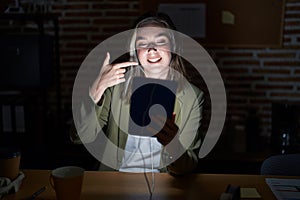 Blonde caucasian woman working at the office at night smiling cheerful showing and pointing with fingers teeth and mouth