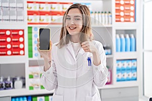 Blonde caucasian woman working at pharmacy drugstore showing smartphone screen smiling happy and positive, thumb up doing