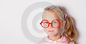 Blonde child with ponytail, in red framed glasses, pink blouse. Smiling, posing with yellow pencil behind ear. Isolated