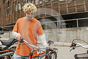 Blonde curly young caucasian guy standing with bike near wooden wall building.