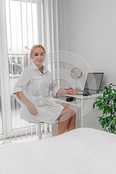 Blonde doctor in the doctor`s office smiling. On the table is a computer, a mirror. The office has white walls, a flower with