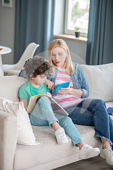 Blonde female and curly boy sitting on sofa, woman helping boy doing his assignment