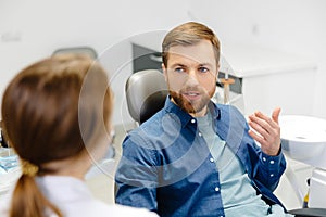 Blonde female dentist in dental office talking with male patient and preparing for treatment. Handsome bearded man in dentist