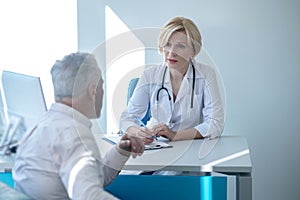 Blonde female doctor sitting at table, writing, consulting gray-haired male patient