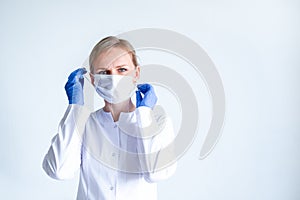 Blonde female doctor surgeon in white uniform, protective blue gloves putting on medical mask over grey. Healthcare