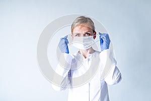 Blonde female doctor surgeon in white uniform, protective blue gloves putting on medical mask over grey. Healthcare