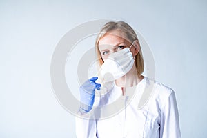 Blonde female doctor surgeon in white uniform, protective blue gloves putting on medical mask over grey. Healthcare