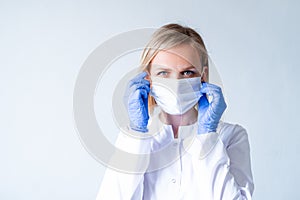 Blonde female doctor surgeon in white uniform, protective blue gloves putting on medical mask over grey. Healthcare