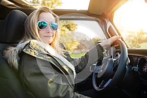 Blonde female driver, driving in nature in convertible with aviator sunglasses and jacket, during sunset