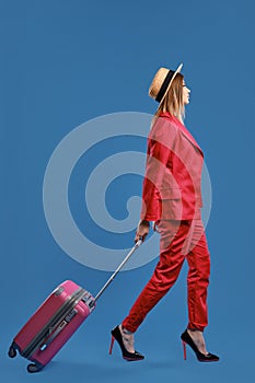 Blonde female in hat, red pantsuit, high heels. She carrying pink suitcase by handle, walking on blue background. Side