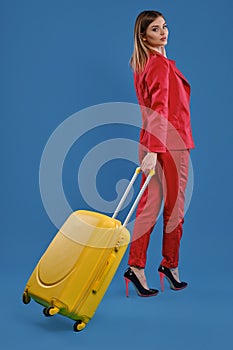 Blonde female in red pantsuit, high black heels. She is posing back to the camera with yellow suitcase on blue studio