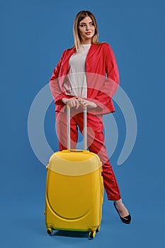 Blonde female in red pantsuit, white blouse and high black heels. She is holding yellow suitcase, posing against blue