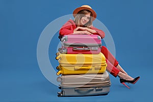 Blonde female in straw hat, red pantsuit, high black heels. Smiling, sitting leaning on three colorful suitcases, posing