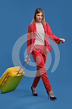 Blonde female in white blouse, red pantsuit and high black heels. She carrying yellow suitcase by handle while walking