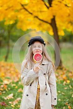 A blonde girl in a beige trench coat and a black cap holds a grapefruit on a stick in her hands