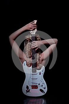 A blonde girl on a black background is sitting with her back and a white guitar