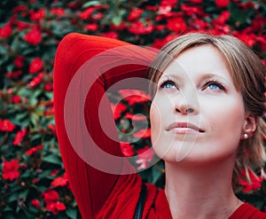 Blonde girl with blue eyes and dressed in red, looks up thoughtful with a background of red flowers