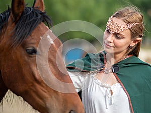 Blonde girl with blue eyes and makeup with elf in the field with a brown horse and a green cape