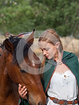 Blonde girl with blue eyes and makeup with elf in the field with a brown horse and a green cape