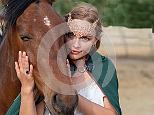 Blonde girl with blue eyes and makeup with elf in the field with a brown horse and a green cape